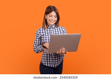 Portrait of successful smiling woman with brown hair typing on laptop keyboard, browsing internet, making presentation, wearing checkered shirt. Indoor studio shot isolated on orange background - Powered by Shutterstock