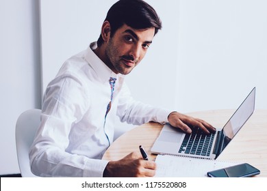 Portrait Of Successful Spanish Entrepreneur Sitting At Table With Desktop Working On Open Laptop Computer And Writing Ideas In Netbook, Young 30 Year Old Businessman Writing Main Theses In Textbook 