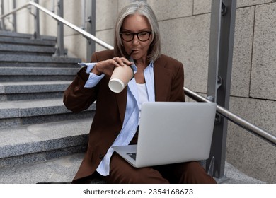 portrait of a successful well-groomed gray-haired middle-aged business woman sitting on the stairs and drinking coffee - Powered by Shutterstock