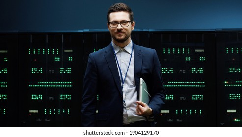 Portrait of successful IT worker with glasses standing in server room folds laptop under one arm and looks into the camera lens.