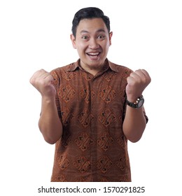 Portrait of successful young Asian man wearing batik shirt shows winning gesture, celebrating victory. Hands raised above, isolated on white