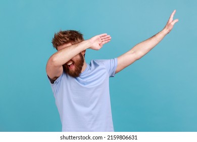 Portrait of successful young bearded man showing dab dance pose, famous internet meme of triumph, performing dabbing trends with hand gesture. Indoor studio shot isolated on blue background.