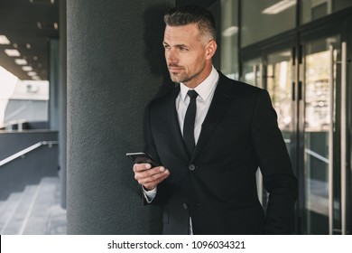 Portrait of a successful young businessman dressed in suit standing outside a glass building and using mobile phone - Shutterstock ID 1096034321