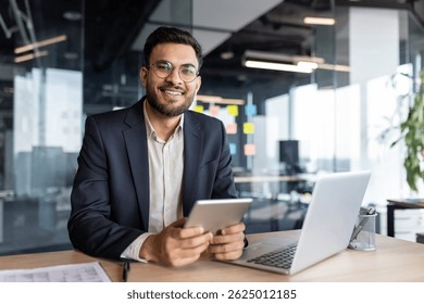 Retrato de un exitoso joven empresario, el hombre está sonriendo y mirando a la cámara. El empleado de la compañía con un traje de negocios sostiene una tableta y una computadora portátil en sus manos.