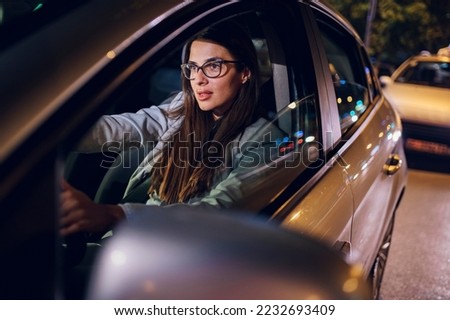 Portrait of a successful young businesswoman in fashion office clothes driving a car in urban modern city during night. People working late. Female holding a steering wheel. Copy space.