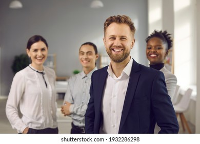 Portrait Of Successful Young Male Business Leader, Company Executive Or Startup CEO With Team Of Happy Employees And Coworkers. Handsome Bearded Caucasian Man In Office Suit Smiling At Camera