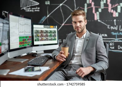 Portrait Of Successful Young Trader Looking At Camera And Drinking Coffee While Sitting In Front Of Multiple Monitors In The Office. Stock Trading, People, Business Concept.