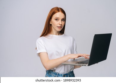 Portrait of successful young woman student holding laptop computer and typing on isolated gray background. Pretty redhead lady model emotionally showing facial expressions in studio, copy space.