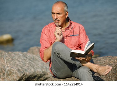 Portrait Of A Sunburnt And Thoughtful Mature Hispanic Man With A Bald Head Looking To The Side Biting A Shackle Of Glasses While Reading A Book Sitting On A Stony Beach Against The Sea. Senior Man