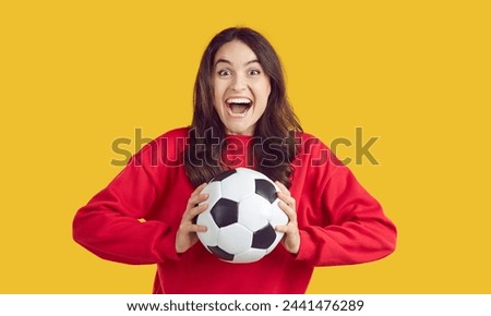 Portrait of super excited beautiful happy brunette woman dressed in warm red jumper holding soccer ball. Studio photo of ardent football fan on isolated yellow background. Concept of sports games.