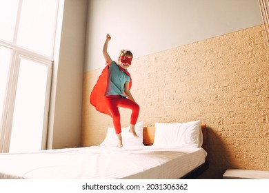 Portrait Of Superhero Kid Jumping On The Bed In Bedroom. Joy And Childhood Concept