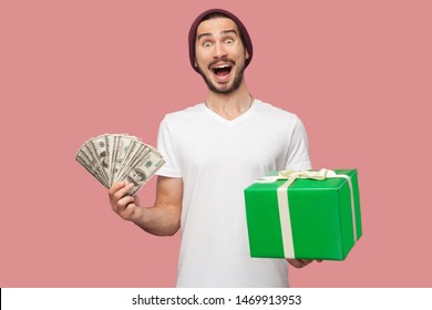 Portrait Of Suprised Handsome Bearded Young Hipster Man In White Shirt And Casual Hat Standing, Holding Fan Of Cash Dollars And Green Present Box. Indoor, Isolated, Studio Shot, Pink Background