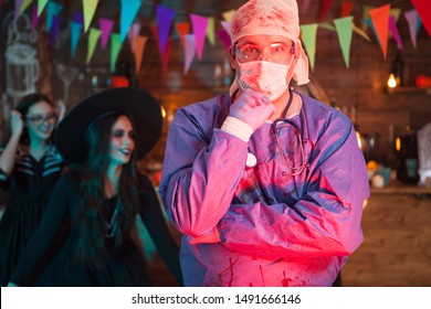 Portrait Of Suprised Man Dressed Up Like A Doctor For Halloween Celebration. Scary Witch In The Background.