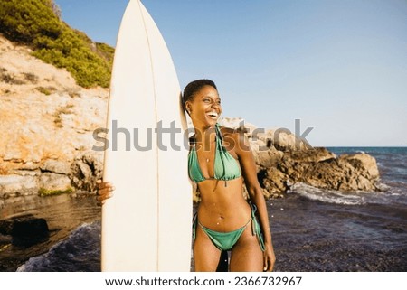 Portrait of a surfer standing on the beach in swimwear, with her surfboard by her side. Happy woman doing beach activities during summer vacation.