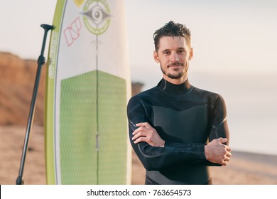 Portrait Of A Surfer With SUP Board On The Beach. Young Man With A Stand Up Paddleboard At Sunset. Extreme Sport Concept. Male Surfer Lifestyle.