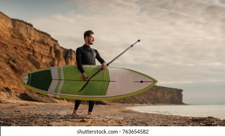 Portrait of a surfer with SUP board on the beach. Young man with a stand up paddleboard at sunset. Extreme sport concept. Male surfer lifestyle.
