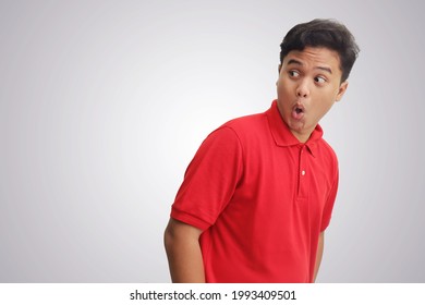 Portrait of surprised Asian man in red polo shirt standing against gray background, showing shocked expression and looking aside