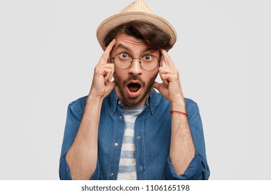 Portrait of surprised bearded young male tries to recollect something very important, has frustrated look and bad memory, wears jean shirt, stylish straw hat, stands against white background