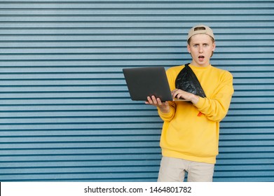 Portrait Of A Surprised Boy Stands With An Open Notebook On A Blue Background, Looks Into A Camera With A Shocking Face, Wears Stylish Streetwear, Cap And A Fanny Pack.