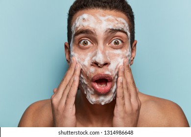 Portrait Of Surprised Brown Eyed Man Washes Face With Cleanser And Soap, Has Foam On Skin, Being Wet, Stares In Mirror, Shocked To Have  Acnes, Isolated Over Blue Background. Men Hygiene