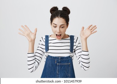 Portrait of surprised brunette girl in striped top and denim overall screaming in excitement, looking down. Cute female is excited with news and gestures actively. Positive emotions and reaction