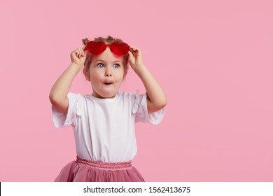 Portrait of surprised cute little toddler girl in the heart shape sunglasses. Child with open mouth having fun isolated over pink background. Looking at camera. Wow funny face