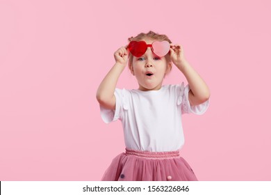 Portrait Of Surprised Cute Little Toddler Girl In The Heart Shape Sunglasses. Child With Open Mouth Having Fun Isolated Over Pink Background. Looking At Camera. Wow Funny Face