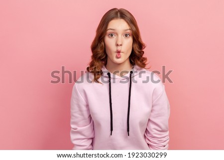 Portrait of surprised funny teen girl with curly hair in hoodie making fish face grimace with pout lips and looking with confused comical expression. Indoor studio shot isolated on pink background