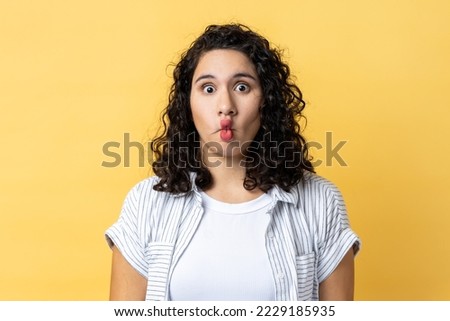 Portrait of surprised funny woman with dark wavy hair making fish face grimace with pout lips and looking with confused comical expression. Indoor studio shot isolated on yellow background.