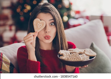 Portrait of surprised girl cover her eye with x-mas ginger bread heart shape cookie wear red sweater in house indoors