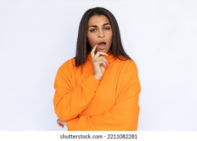 Portrait Of Surprised Hispanic Woman Touching Face. Confused Young Model With Long Hair In Orange Hoodie Looking At Camera With Open Mouth. Studio Shot, Shock Concept.