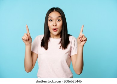 Portrait Of Surprised Korean Woman Gasp In Awe, Pointing Fingers Up, Showing Advertisement, Standing Over Blue Background