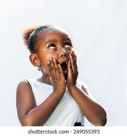 Portrait Of Surprised Little African Girl With Hands On Face Looking Up.Isolated Against Light Background.