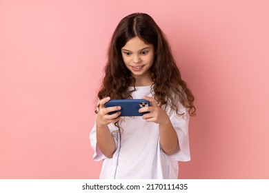 Portrait of surprised little girl wearing white T-shirt using mobile phone with shocked positive expression, playing video game on cellphone. Indoor studio shot isolated on pink background.