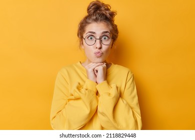 Portrait Of Surprised Lovely Woman Keeps Lips Rounded Hands Under Chin Has Dark Hair Gathered In Bun Wears Round Spectacles And Jumper Isolated Over Yellow Background. Human Face Expressions