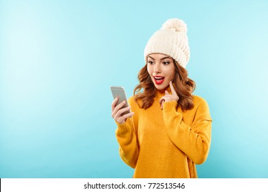 Portrait Of A Surprised Pretty Girl Dressed In Winter Clothes Looking At Mobile Phone Isolated Over Blue Background