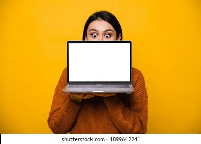Portrait of a surprised shocked caucasian young woman, hiding behind blank white screen laptop, standing on isolated orange background.