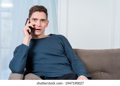 Portrait Of Surprised Shocked Guy, Handsome Young Man With Wide Open Eyes And Mouth Is Talking On Cell Mobile Phone, Calling On Smartphone With Excited Look At Home In Living Room, Sitting At Couch