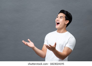 Portrait of surprised smiling young handsome Asian man looking upward and presenting with open palms gesture in isolated studio gray background