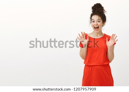Portrait of surprised thrilled beautiful female with curly hair in casual red dress, raising palms and screaming from positive emotions, being impressed and happy while standing against gray wall