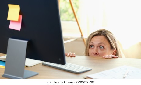 Portrait Of Surprised Woman Peeping Out Table And Looking At Pc Screen. Office Worker Hiding Under Desk