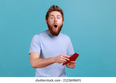 Portrait Of Surprised Young Adult Handsome Bearded Man Reading Letter Or Greeting Card, Holding Envelope, Being Astonished Of Shocked News. Indoor Studio Shot Isolated On Blue Background.