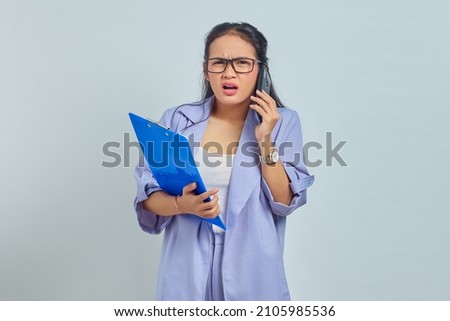 Portrait of surprised young Asian woman laughing while talking on mobile phone with friends about something funny while holding document folder isolated on purple background
