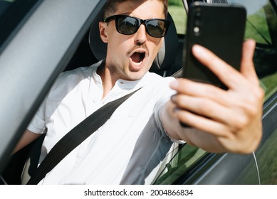 Portrait Of A Surprised Young Man With An Open Mouth Filming An Event On A Mobile Phone While Driving A Car. Selective Focus On The Shocked Face Of The Driver.