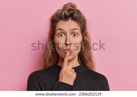 Portrait of surprised young pretty woman keeps lips folded has bugged eyes reacts to something impressive wears casual black jumper isolated over pink background. Human facial expressions concept