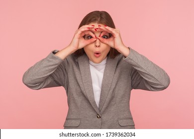 Portrait of surprised young woman in business suit making glasses shape with fingers, looking through binoculars hand gesture and expressing shock, astonishment. indoor studio shot, pink background