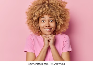 Portrait of surprised young woman with curly bushy hair keeps hands under chin bites lips has wondered expression wears casual t shirt poses against pink background. Human facial expressions - Shutterstock ID 2154049063
