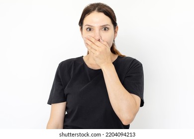 Portrait Of Surprised Young Woman. Female Model In Black T-shirt With Dark Hair Looking At Camera, Covering Mouth With Hand. Portrait, Studio Shot, Surprise Concept