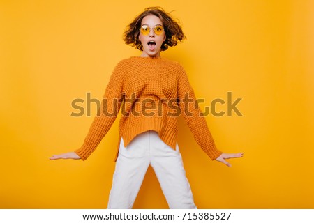 Portrait of surprised young woman in white pants jumping in front of yellow wall. Indoor portrait of curly lady in sunglasses fooling around in studio.