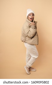 Portrait Of Surprised Young Woman In Winter Coat Pointing Thumb Aside And Looking At Camera Isolated Over Beige Background